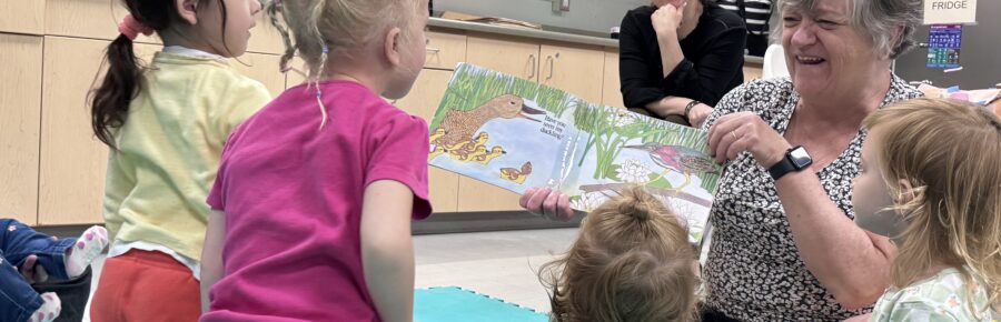 Picture of a group of children excitedly engaging with a book being read and presented by the facilitator, with parents looking on happily in the background.