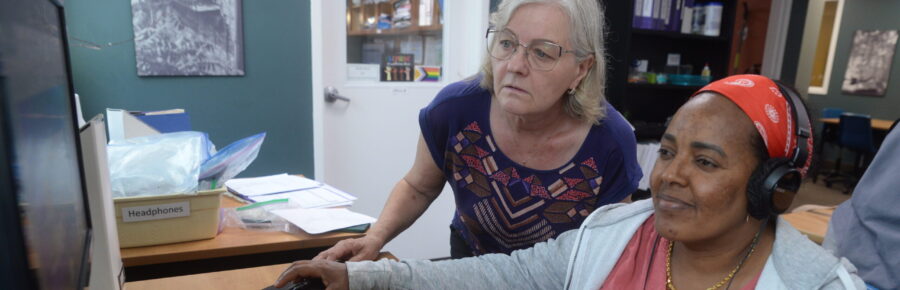 Focused facilitator standing over a smiling learner who is sitting at a desk learning how to use a computer.